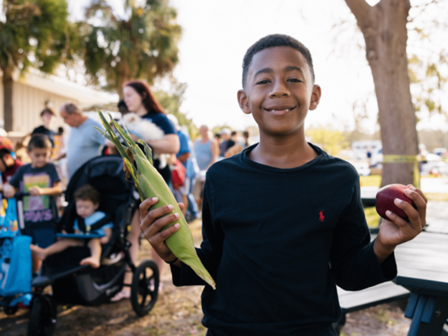 This is a photo from a Feeding America event, features a smiling young boy holding an apple and corn.